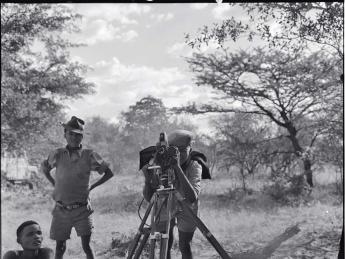 “/Gaishay focusing the camera,” a photograph taken during a 1957-58 ethnographic expedition in Namibia, shows three indigenous boys, one standing behind a camera on a tripod