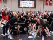 Players shouting with joy surround a large trophy while a fan in the background holds up a giant replica ticket to the NCAA tournament.