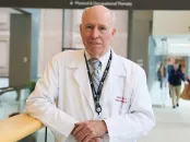 James Muller in white lab coat leaning on railing in hospital hallway.