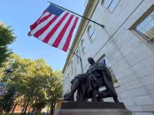 Bronze statue of John Harvard with an American flag in the background, surrounded by trees.