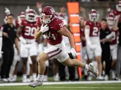 A football player in a maroon jersey sprints past teammates on the sideline.