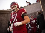 Harvard football player in red jersey 51 walks out of tunnel onto field with teammates behind.