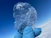 A gloved hand holds a clear shard of blue ice against the sky.