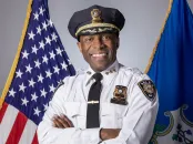 Smiling police chief in uniform, standing with arms crossed between American and state flags.