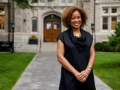 A smiling woman in a black dress stands in front of a stone building on a grassy pathway.