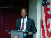 Setti Warren stands at a podium, smiling, with an American flag in the background.