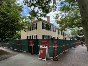 A historic building surrounded by green construction fencing and trees, under a blue sky.