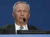 Lawrence Summers in a suit  at a podium with a microphone, looking serious.