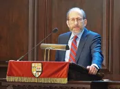 A man in a suit speaking at a podium with a red drape and a university crest.