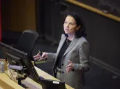 A woman in a gray suit gestures while speaking at a podium in a lecture hall.