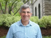 Smiling man in a checkered shirt poses outdoors with greenery in the background.