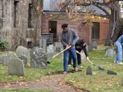 A student uses a shovel to clear debris and weeds from graves in a cemetery