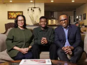 Nancy Hill, Rendall Howell, and their son Theo sit together on a couch in a cozy living room, smiling at the camera.