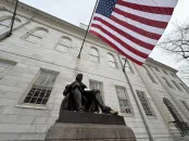 A seated statue of a man reading, with an American flag waving in the background.
