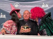 Three people pose together, one holding a small trophy, with playful attire and expressions.