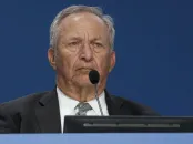 Older man in a suit with a serious expression, seated at a podium with a microphone.