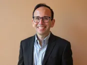 Smiling man in a black suit and glasses, standing against a beige backdrop.