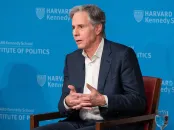 A man in a suit speaks from a chair, with a blue Harvard backdrop.