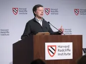 A speaker gestures while presenting at a podium with Harvard Radcliffe Institute branding.