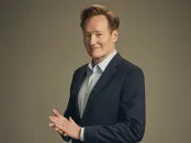 Conan O’Brien in a dark suit and light shirt, smiling and posing with hands clasped.