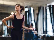 A dancer in a black leotard poses gracefully in a bright studio, with mirrors reflecting her movement.