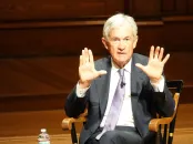 A man with gray hair gestures with both hands while seated on a wooden stage.