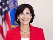 Rochelle Walensky in a red blazer with short dark hair, standing in front of flags.