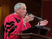 Former Harvard President Lawrence S. Bacow in a red academic robe gestures passionately at a podium, with seated audience members behind.