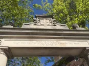 Stone archway adorned with a crest and surrounded by green trees.