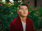 Man in a red jacket sitting against a backdrop of green foliage and pink flowers.