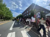 Protesters holding signs march along a sunny street in front of a building.