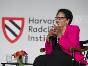 A woman in a pink blazer smiles while seated on stage at a Harvard event.