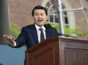 Ronny Chieng wearin a suit speaks passionately at a wooden podium outdoors.