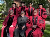 Group of seven individuals in academic regalia, smiling outside on a sunny day.