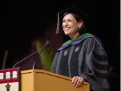 A smiling woman in academic regalia speaks at a podium during a graduation ceremony.