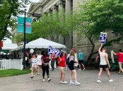 People walking with signs at an outdoor event, near a tent and greenery.