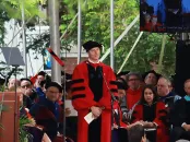 A speaker in academic regalia stands before an audience during a ceremony outdoors.