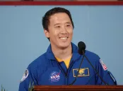 Smiling astronaut in blue jumpsuit speaking at a podium, colorful background.