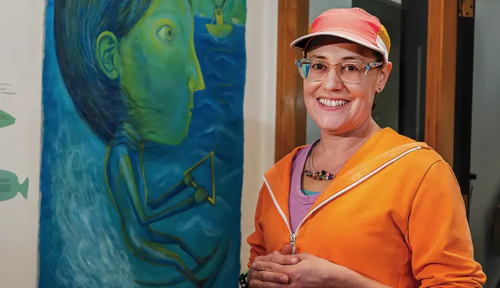 A woman in an orange sweater and cap stands in front of a large blue and green painting of a water-skiing woman.