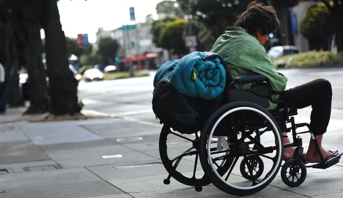 A man in a wheelchair with a rolled-up sleeping bag strapped to the back