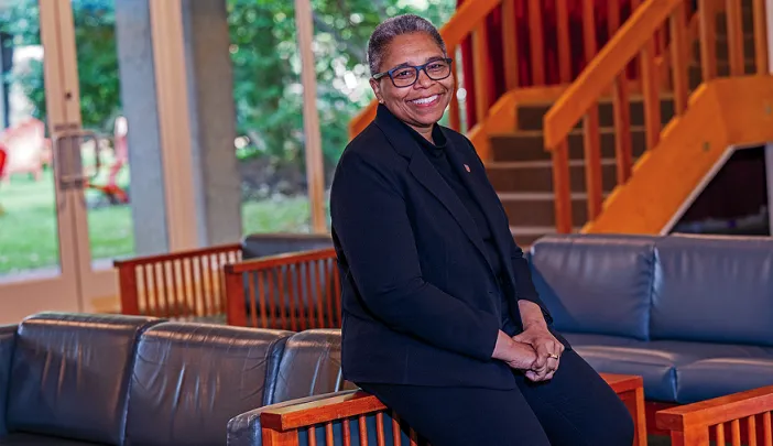 Smiling woman in glasses and a dark outfit seated indoors near a staircase and large windows.
