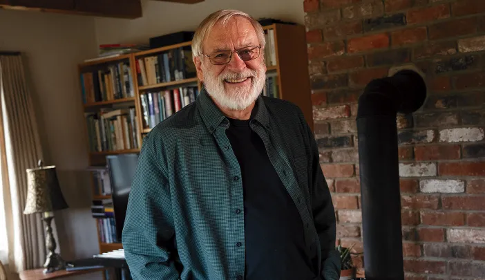 Tom Lee standing in his home in New Hampshire, surrounded by books and a brick fireplace.