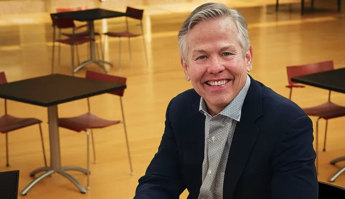 Tom Kane smiles  in a navy blazer and sits at a table in a modern room with empty chairs and tables behind him.