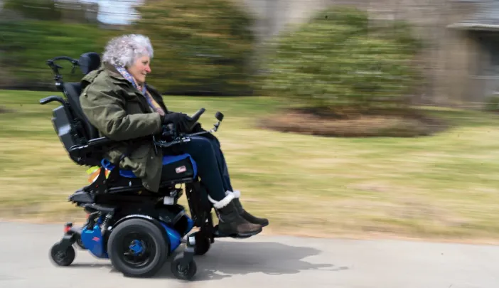 Lisa Iezzoni smiling while riding a motorized wheelchair on a paved path through a grassy area