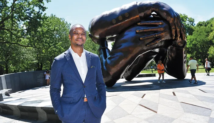 Brandon Terry, wearing a blue suit, standing before The Embrace, a large bronze sculpture of intertwined arms in Boston Common.