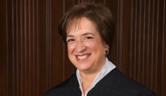 Justice Elena Kagan in a black robe, smiling against a wooden panel background.