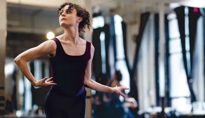A dancer in a black leotard poses gracefully in a bright studio, with mirrors reflecting her movement.