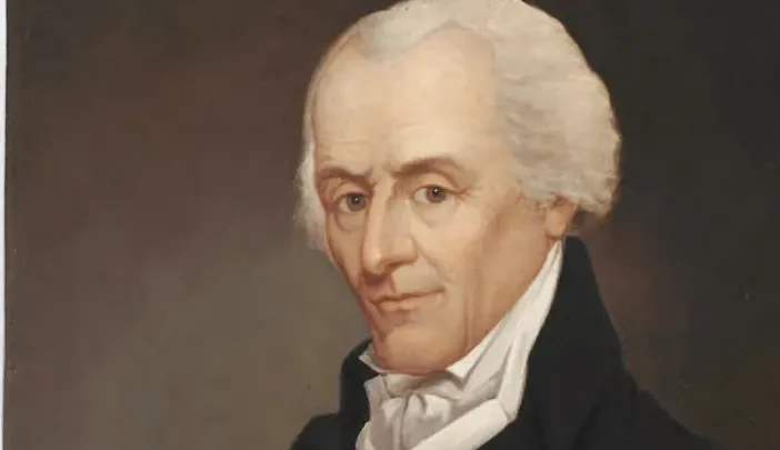 Portrait of a man with white hair, wearing a black coat, arms crossed, thoughtful expression.