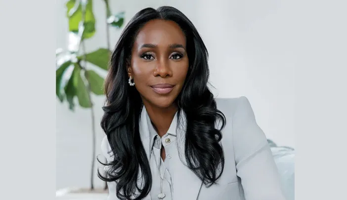 A woman with long, wavy hair wearing a white suit, seated in a bright, modern room.