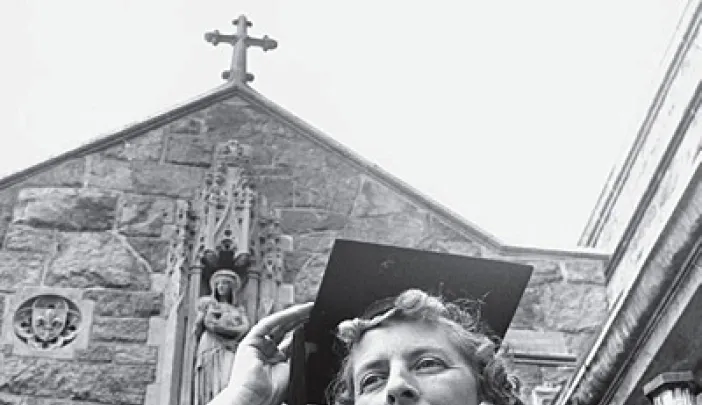 Woman in graduation cap smiling outdoors, church in the background, black and white photo.